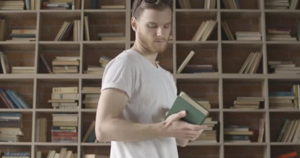 若いブルネットの男は本棚からカメラに向かい眼鏡をかける。図書館で本を読んでいるハンサムな髭の白人男性の肖像画。知性の概念だ。シネマ4k｜ProRes HQ. — ストック動画