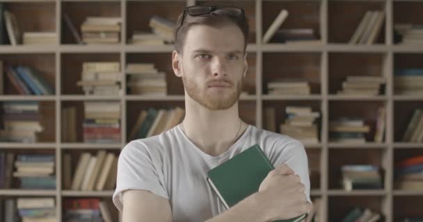 Przemyślany, przystojny młodzieniec stojący w bibliotece z książką w ręku i patrzący w kamerę. Portret poważnej białoruskiej dziennikarki, autora lub biznesmena. Styl życia. Kino 4k ProRes kwatera główna. — Wideo stockowe