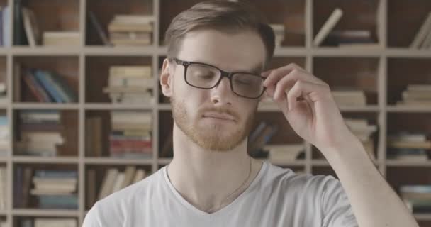 Close-up portret van een man die een bril op het hoofd legt en handen kruist. Jonge knappe blanke man die poseert in de bibliotheek op de achtergrond van boekenplanken. Cinema 4k ProRes Hoofdkwartier. — Stockvideo