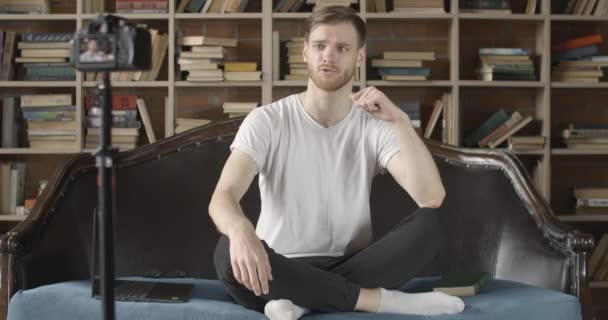 Blogueiro milenar positivo gravando vídeo sobre literatura na biblioteca. Retrato do jovem caucasiano bonito conversando na câmera com estantes de livros no fundo. Vlogagem. Cinema 4k ProRes HQ . — Vídeo de Stock