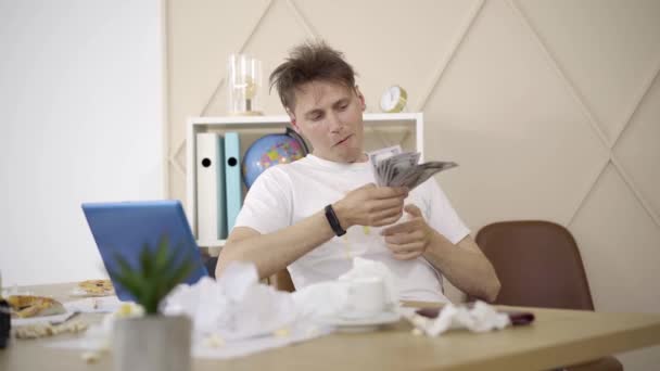 Retrato de un joven contando dinero sentado a la mesa en una habitación desordenada. Morena chico caucásico en camiseta sucia que tiene buenos ingresos trabajando desde casa. Trabajo remoto, estilo de vida independiente . — Vídeo de stock