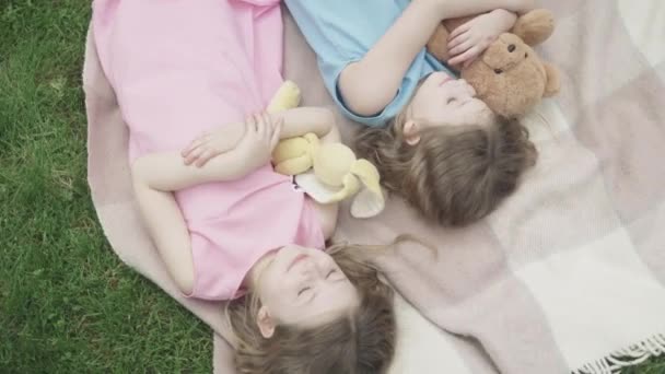 카메라는 야외에서 담요 위에서 장난감을 가지고 자는 귀여운 소녀들 주변을 돌아다니고 있다. 눈을 감은 채푸른 초원에 누워 있는 아름다운 카프카스 쌍둥이 자매들의 맨 위에 있는 모습. 여가, 휴식, 즐거움. — 비디오