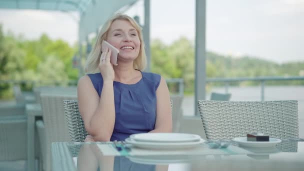 Porträt einer positiven erwachsenen kaukasischen Frau, die am Telefon wie am Restauranttisch im Freien sitzt. Selbstbewusste, charmante blonde Dame in blauem Kleid, die im Café am Flussufer ruht. Lebensstil. — Stockvideo