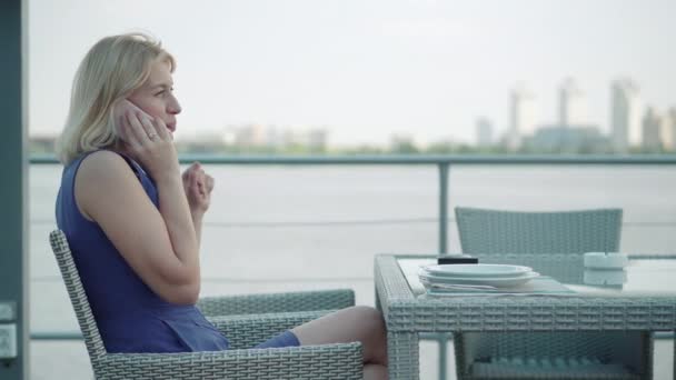 Seitenansicht der schönen kaukasischen Frau, die am Telefon wie im Restaurant im Freien sitzt. Porträt einer eleganten, selbstbewussten blonden Dame in blauem Kleid, die in einem Café am Ufer des Flusses ruht. Lebensstil. — Stockvideo