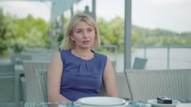 Zarif mavi elbiseli üzgün, yalnız beyaz kadın açık hava restoranında oturuyor ve başka tarafa bakıyor. Nehir kıyısındaki kafede tek başına dinlenen büyüleyici yetişkin kadın turistin portresi. Yalnızlık, yaşam tarzı.