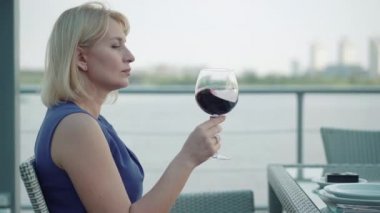 Yan görüntüde başarılı iş kadını kırmızı şarabı sallıyor, kokluyor ve tadıyor. Açık hava restoranında içki içen zarif beyaz kadın portresi. Boş zaman, güzellik, lüks, yaşam tarzı.