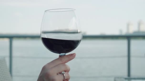 Copa de vino tinto en mano femenina caucásica en el fondo del tranquilo río urbano. Mujer irreconocible sacudiendo el vino y sonriendo. Señora disfrutando de la bebida en el restaurante al aire libre. lujo, estilo de vida . — Vídeos de Stock