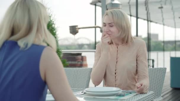 Jovencita alegre hablando con una amiga irreconocible en el café. Retrato de damas rubias emocionadas charlando en un restaurante al aire libre en la orilla del río. Amistad, ocio, estilo de vida, chismes . — Vídeos de Stock