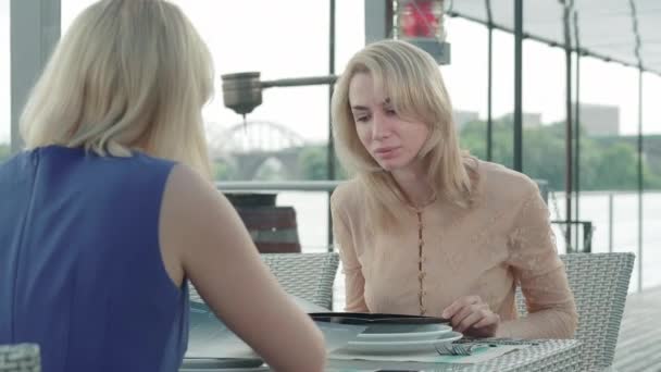 Retrato de una mujer rubia concentrada sentada con un amigo en el café y discutiendo el menú. Mujeres amigas caucásicas descansando en un restaurante al aire libre y hablando. Alegría, ocio, estilo de vida . — Vídeos de Stock