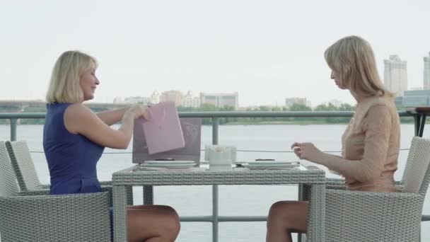 Mujer caucásica feliz dando regalo para una amiga en un restaurante al aire libre. Vista lateral amplia toma de mujeres positivas que se reúnen en la cafetería para la celebración. Alegría, estilo de vida, ocio, vacaciones . — Vídeos de Stock