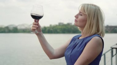 Açık kafede gülümseyen sarışın, beyaz bir kadının kırmızı şarapla bardağı sallamasının yan görüntüsü. Restorandaki nehir kıyısında içki içen kendine güvenen güzel bir bayan. Zarafet, alkol endüstrisi, yaşam tarzı.