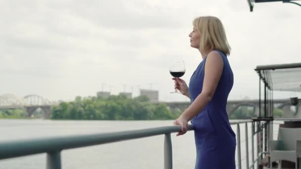 自信を持って思慮深い女性がワイングラスと川の銀行の上に立って離れて見ての側のビュー。ウォーターフロントのレストランで休んで魅力的なエレガントな白人女性の肖像画。自信と富. — ストック動画