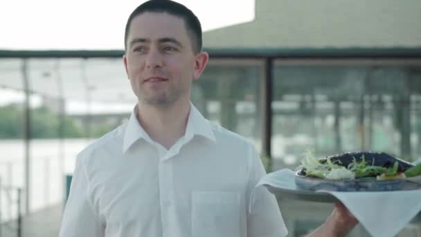 Portrét pozitivního kavkazského číšníka kráčejícího s jídlem ve venkovní restauraci. Profesionální zaměstnanec podávající jídlo v kavárně venku. Důvěra, povolání, životní styl, zaměstnání, zaměstnání. — Stock video