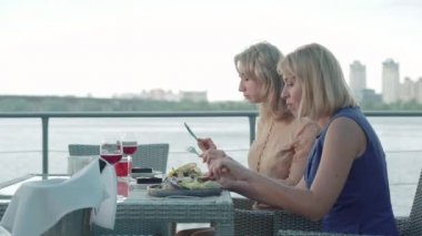 Açık hava restoranında yemek yiyen Kafkasyalı zengin kadınların yan görüntüsü. Masada kırmızı şarap ve yemeklerle konuşan ve lezzetli yemekler yiyen kendine güvenen kadınların portresi. Yaşam tarzı, zenginlik..