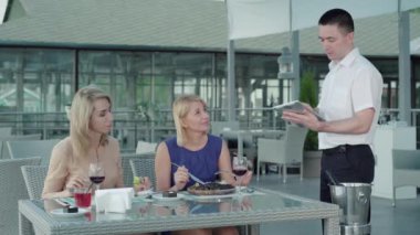 Ciddi bir beyaz adam açık hava restoranında sipariş veriyor. Kafede zengin bayan müşterilere servis yapan profesyonel erkek garsonun portresi. Sarışın kadınlar yemek sipariş ediyor. Yaşam tarzı, rahatlık, beslenme.