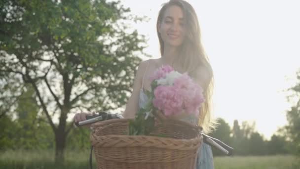 Vacker brunett kvinna tar bukett från cykelkorg och luktar blommor. Porträtt av en elegant vit flicka i solstrålar vid solnedgången. Charmig dam vilar i sommarparken vid solnedgången. — Stockvideo