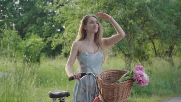 Tiro médio de mulher morena elegante em pé no parque de verão com bicicleta e olhando para longe. Jovem menina branca sorridente de mão dada na testa. Bela senhora positiva desfrutando fins de semana ao ar livre . — Vídeo de Stock