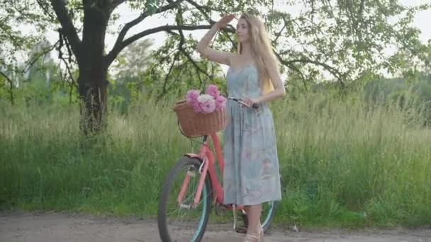 Tânăra femeie caucaziană subțire cu bicicleta uitându-se departe ținându-se de mână la frunte. Portret mare împușcat de fermecător fericit bruneta doamnă în rochie bucurându-se de apus de soare în parc în aer liber. Bucurie, petrecere a timpului liber, stil de viață . — Videoclip de stoc