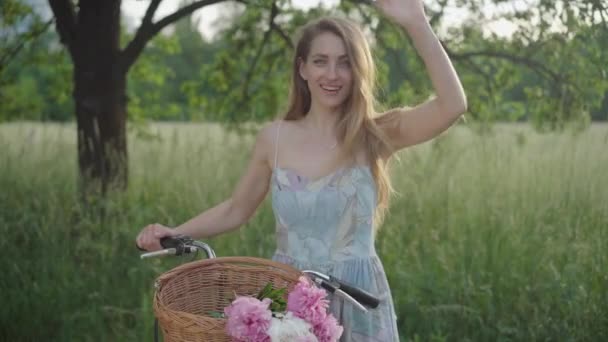 Retrato plano medio de la encantadora mujer sonriente saludando de la mano como de pie con bicicleta en el parque de verano. Atractiva chica caucásica en vestido retro posando con bicicleta al aire libre. Ocio, belleza . — Vídeos de Stock