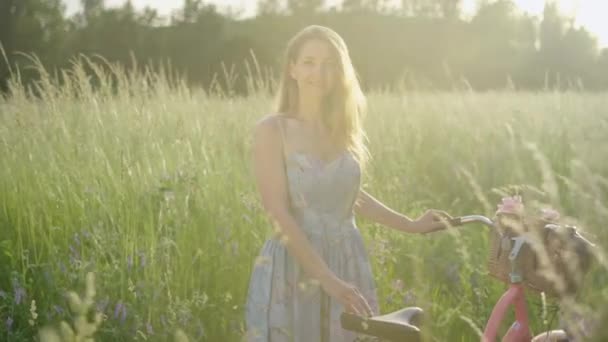 Retrato de una encantadora joven posando con bicicleta en el soleado prado. Joven dama caucásica sonriente en vestido de pie en la luz del sol al aire libre y sonriendo a la cámara. Belleza, estilo retro . — Vídeos de Stock