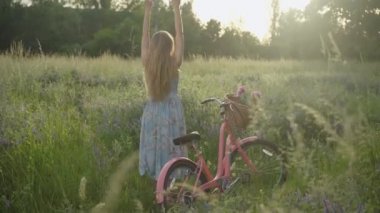 Gün batımında kollarını kaldırmış genç beyaz kadının geniş açılı görüntüsü. Çiçekler sepette, çayırda zarif retro elbiseli kadın bisikletçinin yanında duran bisiklet..