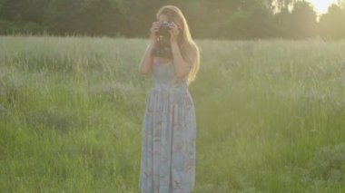 Geniş açıda, genç bir kadın kamerayla güneşli bir çayırda fotoğraf çekiyor. Çekici beyaz kadın fotoğrafçının portresi gün batımında dışarıda hobi yapıyor..