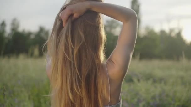 На задньому плані жінка регулює волосся і піднімає руки на сонці. Чарівна струнка молода кавказька дівчина, яка насолоджується сонячним літнім вечором у парку на лузі. Право, спокій, щастя. — стокове відео