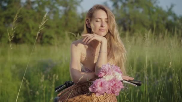 Retrato de menina elegante posando com buquê de flores em cesta de bicicleta. Mulher branca morena positiva olhando para a câmera e sorrindo como estando no ensolarado prado de verão no parque . — Vídeo de Stock