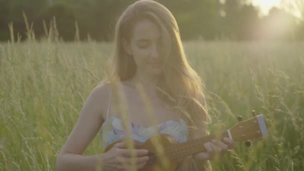 Imagem do meio da jovem morena tocando ukulele ao pôr do sol no parque e sorrindo para a câmera. Retrato de bela mulher caucasiana encantadora descansando ao pôr-do-sol no prado. Conceito de beleza natural . — Vídeo de Stock