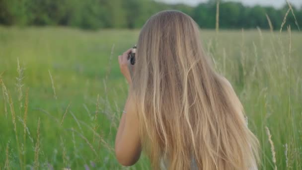 Vista posterior de una joven fotógrafa tomando fotos del paisaje en el soleado día de verano. Mujer caucásica profesional delgada fotografiando la naturaleza en el prado en el parque. Estilo de vida, hobby . — Vídeos de Stock