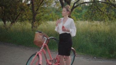 Yaz parkında bisikletle duran ve meyve suyu içen güzel, yorgun iş kadını. Resmi iş kıyafetlerinde gün batımında mola veren beyaz kadın portresi. Neşe, yaşam tarzı.