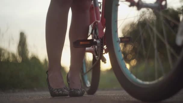 Primer plano de las piernas femeninas en tacones altos de pie junto a la bicicleta al atardecer. Mujer caucásica irreconocible descansando en el callejón del parque de verano. Ocio, relajación, estilo de vida saludable . — Vídeos de Stock