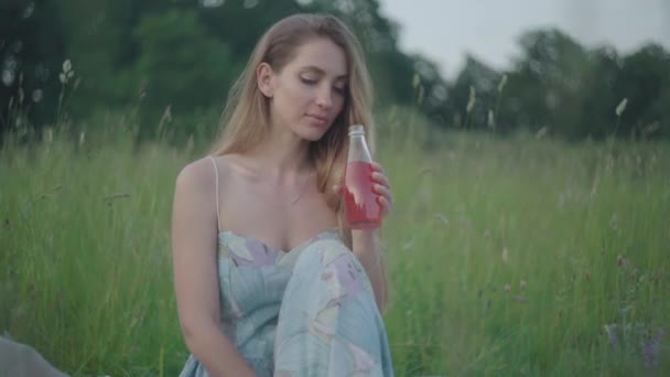 一个积极的年轻女子在夏天的草地上喝果汁的画像。迷人的白种人女孩穿着雅致的衣服在户外享受周末.休闲、快乐、生活方式、快乐. — 图库视频影像