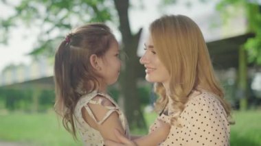 Yan görüntüde gülümseyen sarışın anne ve esmer küçük kızın parkta sarılıp konuştuklarını görüyoruz. Neşeli Kafkasyalı ebeveyn ve çocuğun mutlu yüzleri açık havada güneşli bir yaz gününün tadını çıkarıyor..