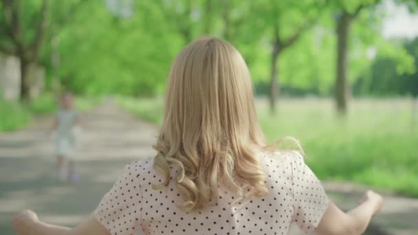 Vista posteriore della donna bionda caucasica seduta sul vicolo nel parco estivo mentre i bambini corrono da lei. Figlia allegra che abbraccia la madre, figlio che passa davanti. Famiglia godendo giornata di sole all'aperto. — Video Stock