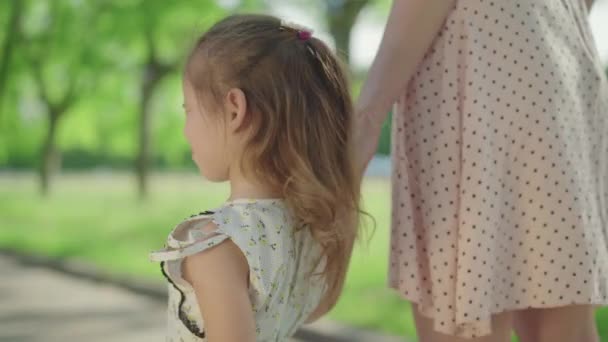 Linda chica sonriente girando a la cámara y sonriendo. Retrato de la encantadora hijita caucásica cogida de la mano de la madre en el soleado parque de verano. Alegría, ocio, infancia, felicidad . — Vídeos de Stock