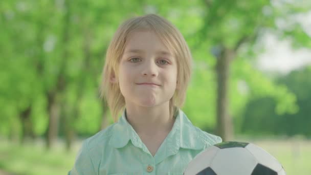Primer plano del encantador chico rubio caucásico con ojos verdes posando con pelota de fútbol en el soleado parque. Retrato de un niño feliz sonriendo a la cámara disfrutando del día de verano al aire libre. Infancia, hobby, fútbol . — Vídeo de stock