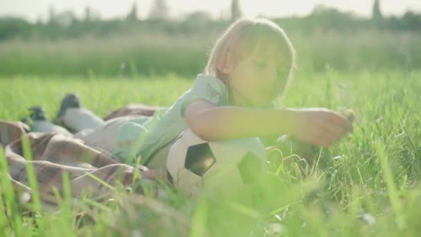 Porträt eines charmanten kleinen Jungen, der den sonnigen Sommertag auf der Wiese genießt. Nettes kaukasisches blondes Kind, das mit Fußball auf grünem Gras liegt und in die Kamera schaut. Freizeit, Kindheit, Erholung. — Stockvideo