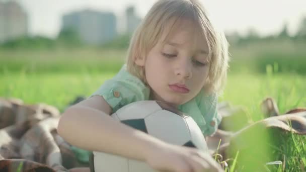 Primer plano de niño rubio reflexivo con ojos verdes que yacen en el prado verde con pelota de fútbol. Retrato de un lindo niño caucásico descansando en un día soleado al aire libre. Estilo de vida, infancia . — Vídeos de Stock