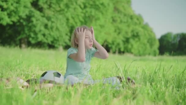 Trauriger blonder Kaukasier, der auf einer grünen Wiese sitzt und den Kopf mit den Händen hält. Weitwinkelporträt eines aufgebrachten kleinen Kindes mit Fußballball, das einen sonnigen Sommertag allein im Freien verbringt. — Stockvideo