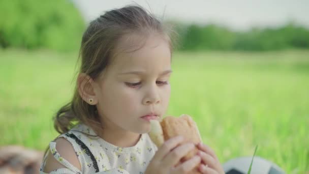 ใกล้ชิดของสาวสีน้ําตาลน้อยน่ารักที่มีตาสีน้ําตาลกัดครัวซองต์ โฟกัสเปลี่ยนจากใบหน้าที่สวยงามของเด็ก Caucasian ไปเป็นขนมปังอบอร่อย เด็กน่ารักพักผ่อนบนทุ่งหญ้าสีเขียวในวันฤดูร้อน . — วีดีโอสต็อก