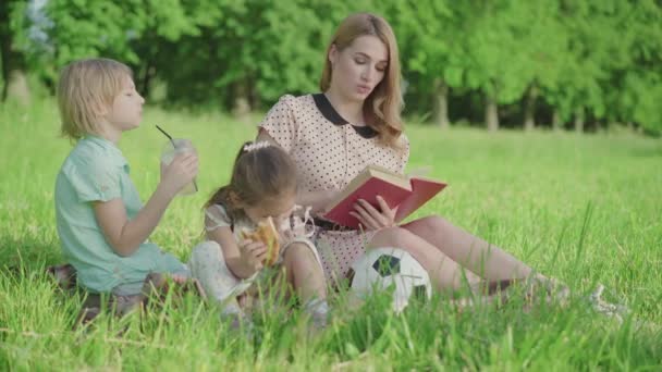 Portret atrakcyjnej młodej matki czytającej książki dla dzieci na zielonej letniej łące. Szczęśliwa biała kobieta odpoczywa z synem i córką na świeżym powietrzu w słoneczny dzień. Wypoczynek, rodzina, styl życia. — Wideo stockowe