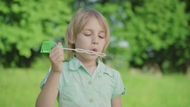 Close-up de menino caucasiano soprando bolhas de sabão no prado ensolarado. Criança loira bonito com olhos verdes descansando ao ar livre no dia de verão. Lazer, alegria, infância, relaxamento . — Vídeo de Stock