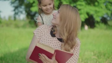 Sevimli küçük bir kız annesine gelip parkta kitap okuyor ve gözlerini kapıyor. Güzel kızıyla güneşli hafta sonlarının tadını çıkaran pozitif beyaz kadın portresi. Annelik, çocukluk, yaşam tarzı.