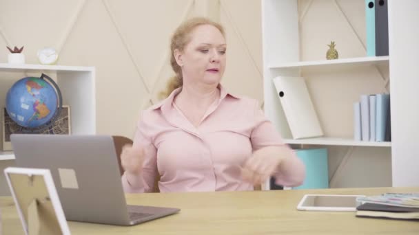 Sıcaktan bunalmış yaşlı bir kadının portresi. Masada dizüstü bilgisayarla oturan beyaz bir kadın yelpaze sallıyor. İklim değişiklikleri, menopoz, kadın yaşlanma konsepti. — Stok video