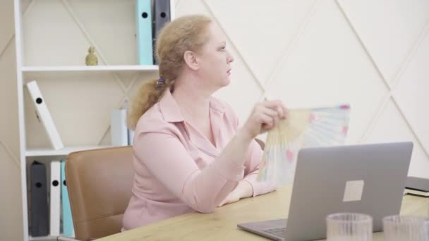 Vista laterale della donna anziana nervosa che agita il ventilatore e tiene la testa con mano. Ritratto di donna caucasica anziana che suda al chiuso. Concetto di menopausa, cambiamenti climatici, invecchiamento femminile. — Video Stock