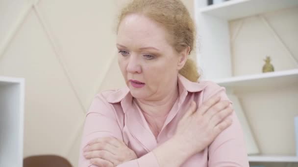 Nahaufnahme einer Seniorin, die sich beim Aufwärmen die Schultern reibt. Rothaarige kaukasische Dame, die sich drinnen kalt fühlt. Unzufriedene Frau sitzt am Tisch und blickt auf Laptop-Bildschirm. — Stockvideo