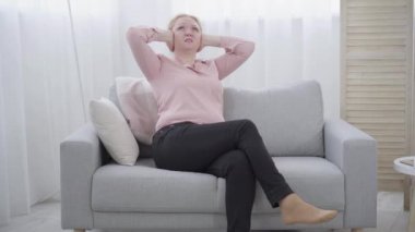 Mutsuz yaşlı kadın, elleri kulakları kapalı bir şekilde koltukta oturuyor. Komşuların gürültüsünden muzdarip beyaz kadının geniş açılı portresi. Konut sorunları, yaşam tarzı.