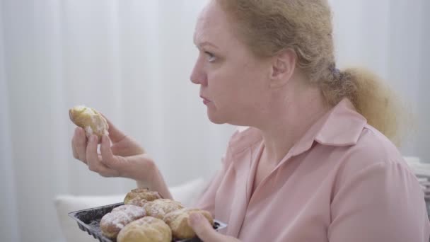 Close-up vista lateral da mulher gordinha sênior mastigar pão doce. Retrato de mulher ruiva caucasiana com obesidade a comer padaria pouco saudável. Comida de plástico, gordura . — Vídeo de Stock