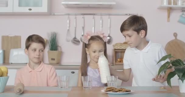 Retrato de um cavalheiro a derramar leite em copos na cozinha. Três crianças sentadas à mesa em casa a almoçar. Meninos e meninas gêmeos descansando juntos dentro de casa. Conceito de amizade . — Vídeo de Stock